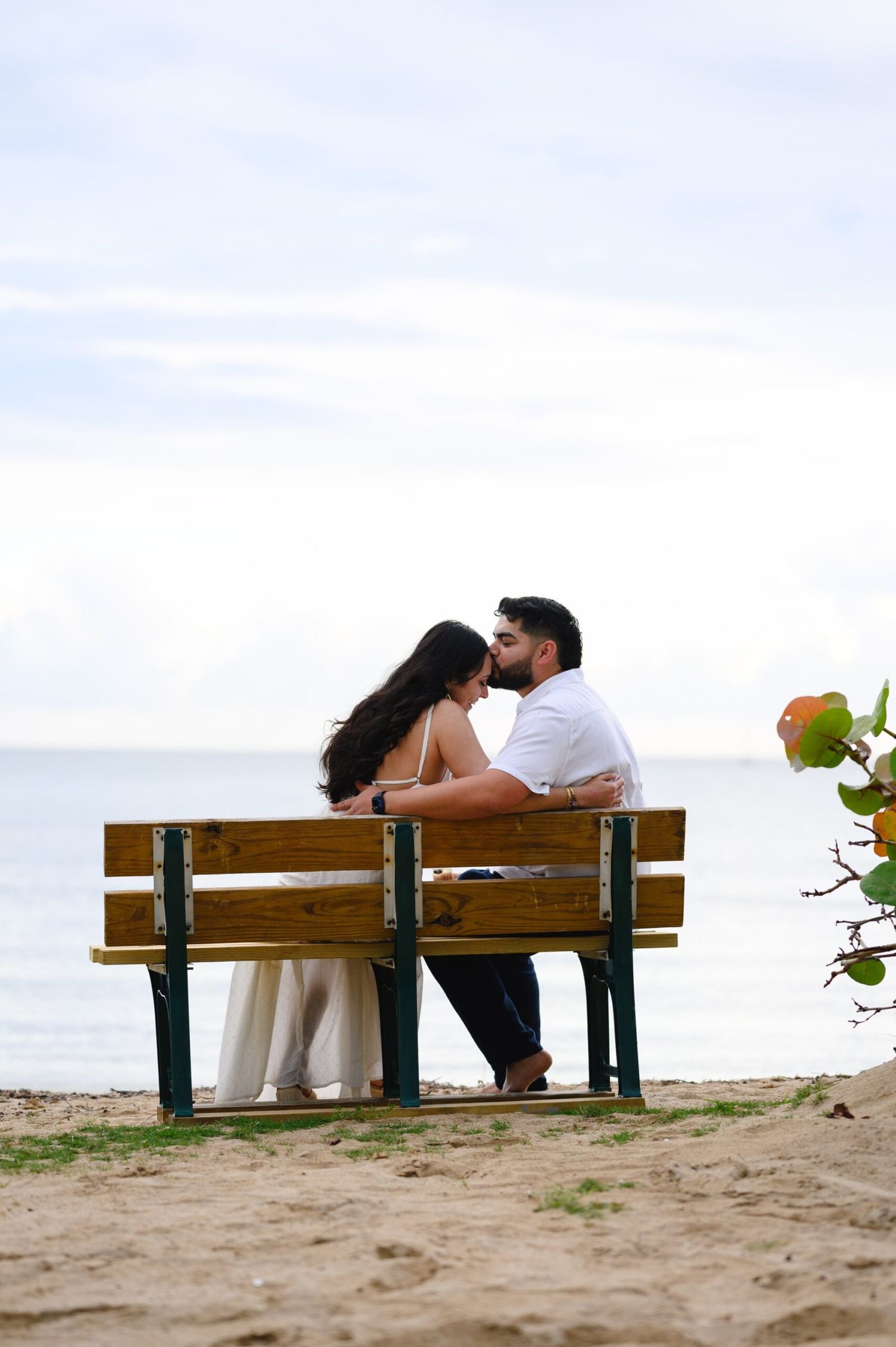 Couple sitting together on a wooden bench by the beach, sharing a romantic moment with the ocean in the background.