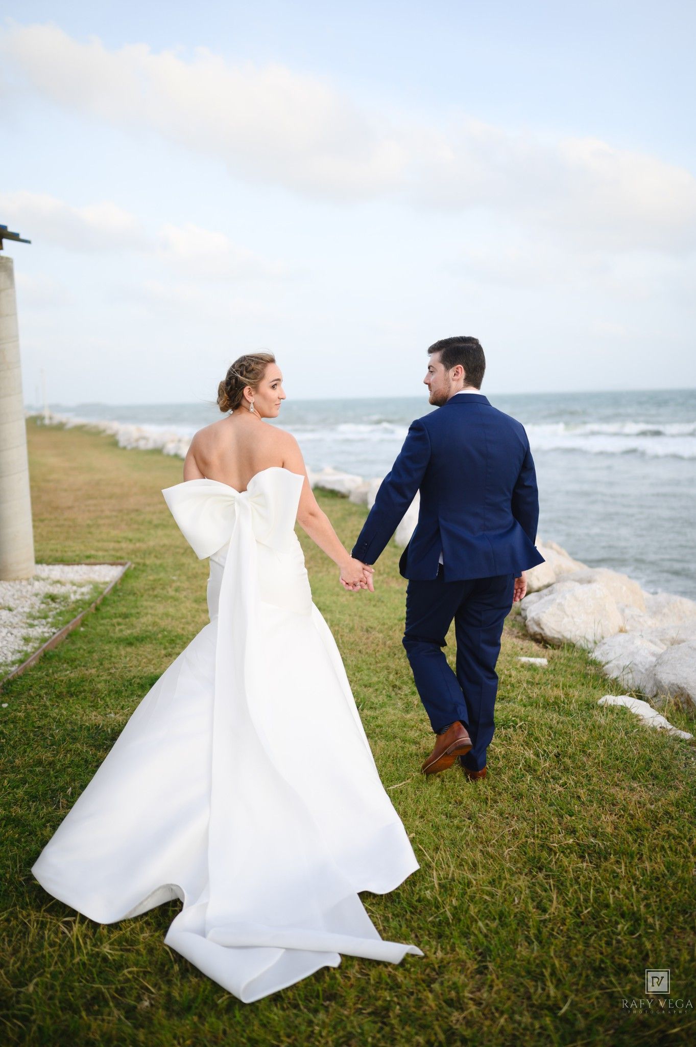 Bride in white off-shoulder gown with bow and groom in navy suit walk hand-in-hand along oceanside grass path.