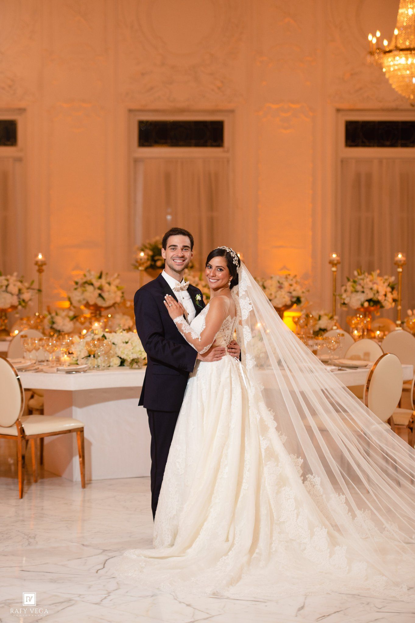 Bride and groom pose elegantly in a grand ballroom with golden lighting, floral centerpieces, and a flowing cathedral veil.