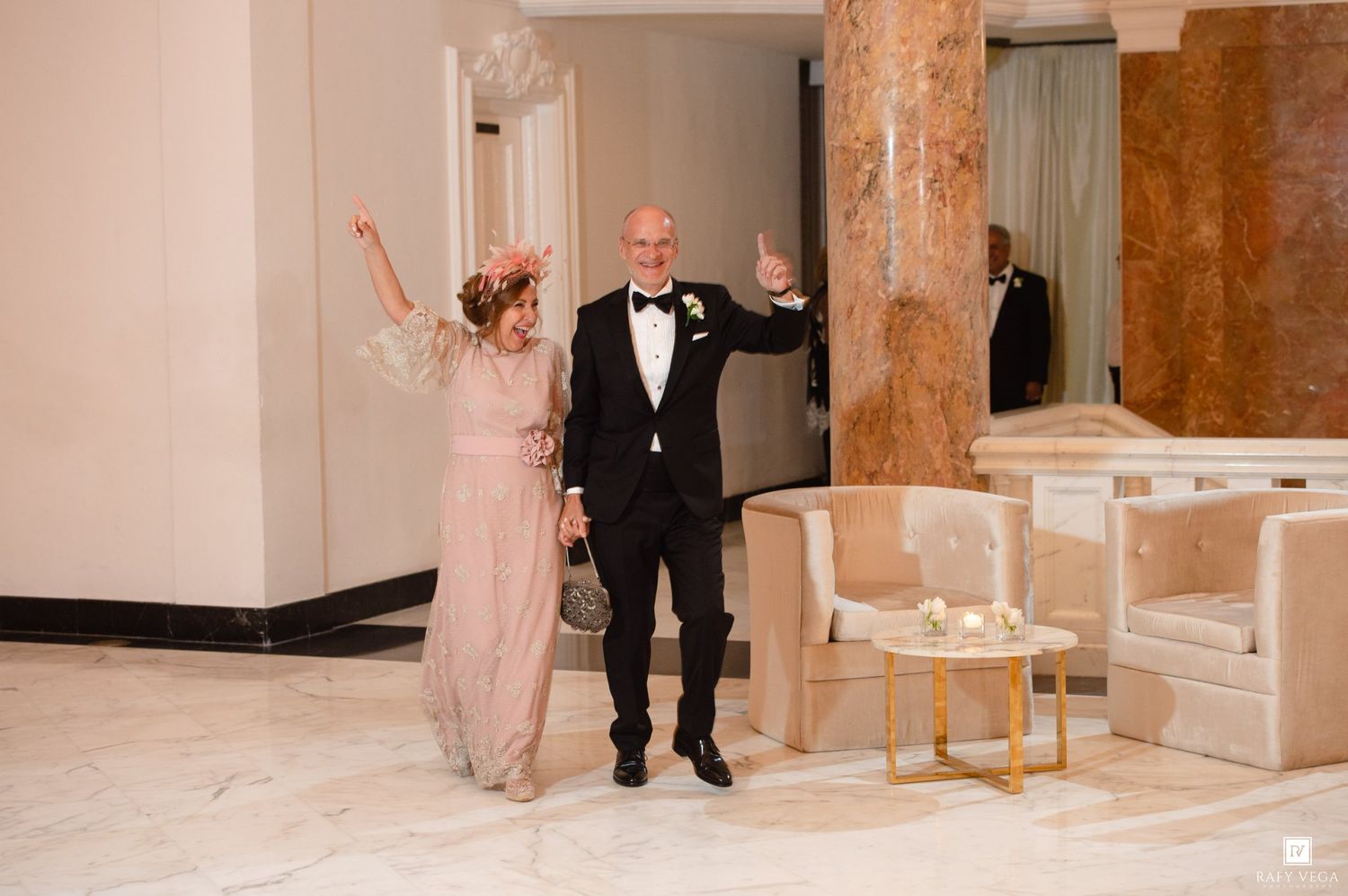 Elegant couple in formal attire celebrating in a luxurious marble lobby, woman in pink gown and man in tuxedo.
