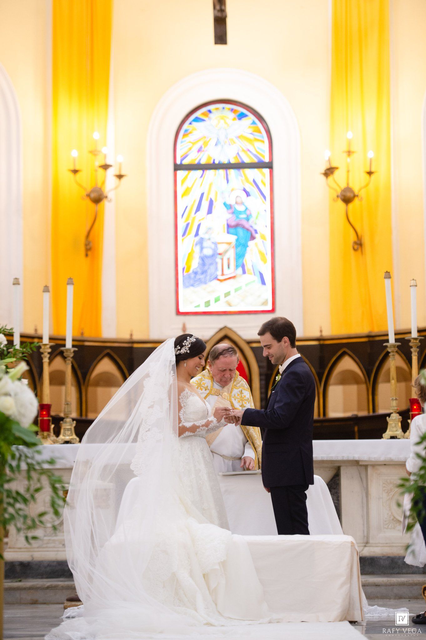 Bride and groom exchange vows at church altar with stained glass window and candles during wedding ceremony.