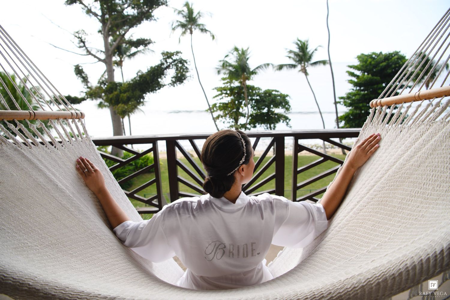 Bride in white robe relaxing in hammock on tropical resort balcony overlooking lush greenery and ocean.