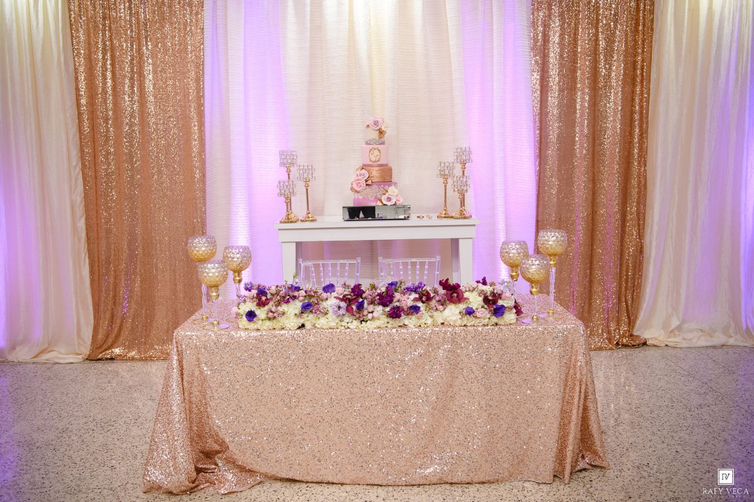 Elegant wedding reception with gold sequin tablecloth, floral arrangements, pink cake, and draped backdrop.