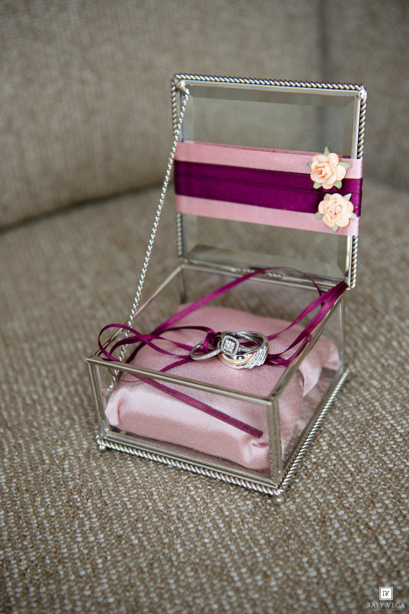 Glass jewelry box with pink cushion, purple ribbon, and wedding rings on a gray sofa.