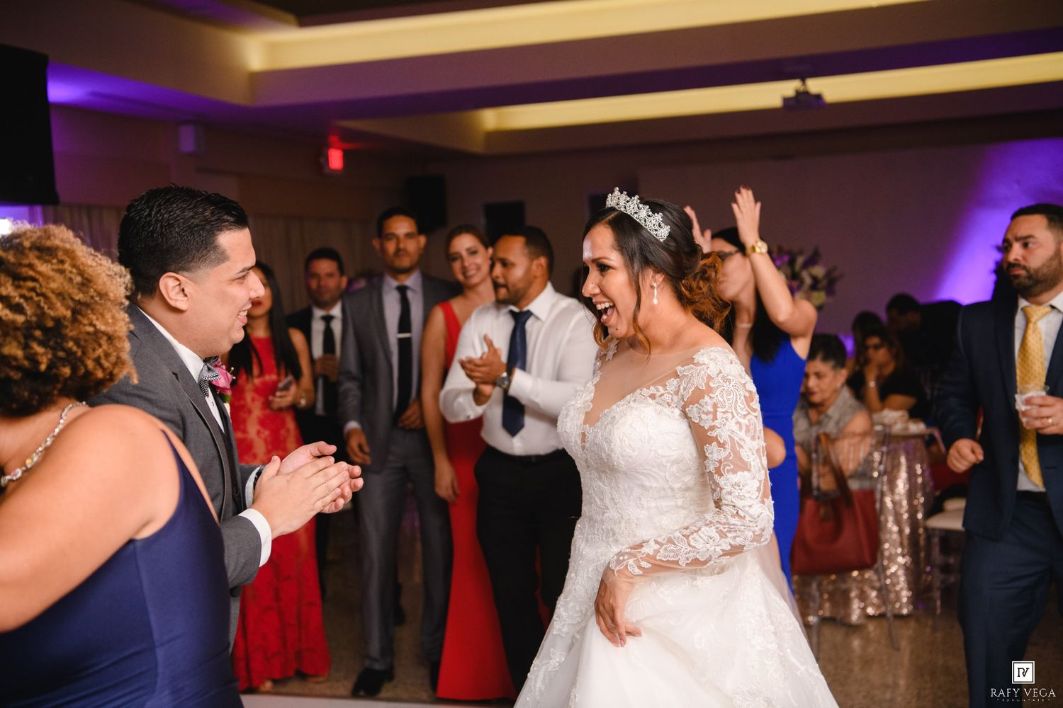 Bride in lace wedding gown dancing joyfully with guests at elegant wedding reception with purple lighting.