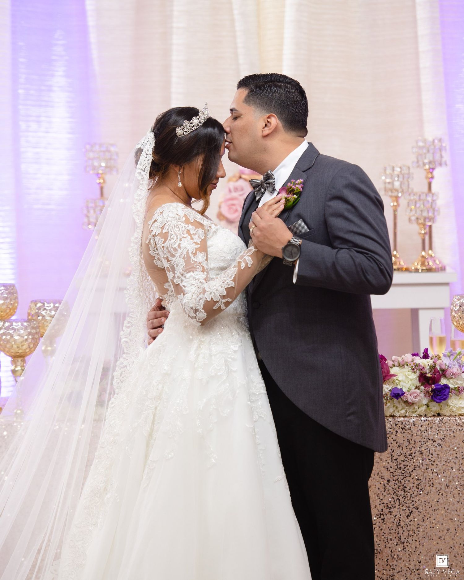 Bride and groom sharing a kiss at their elegant wedding reception, bride wearing lace gown with veil, groom in gray suit.