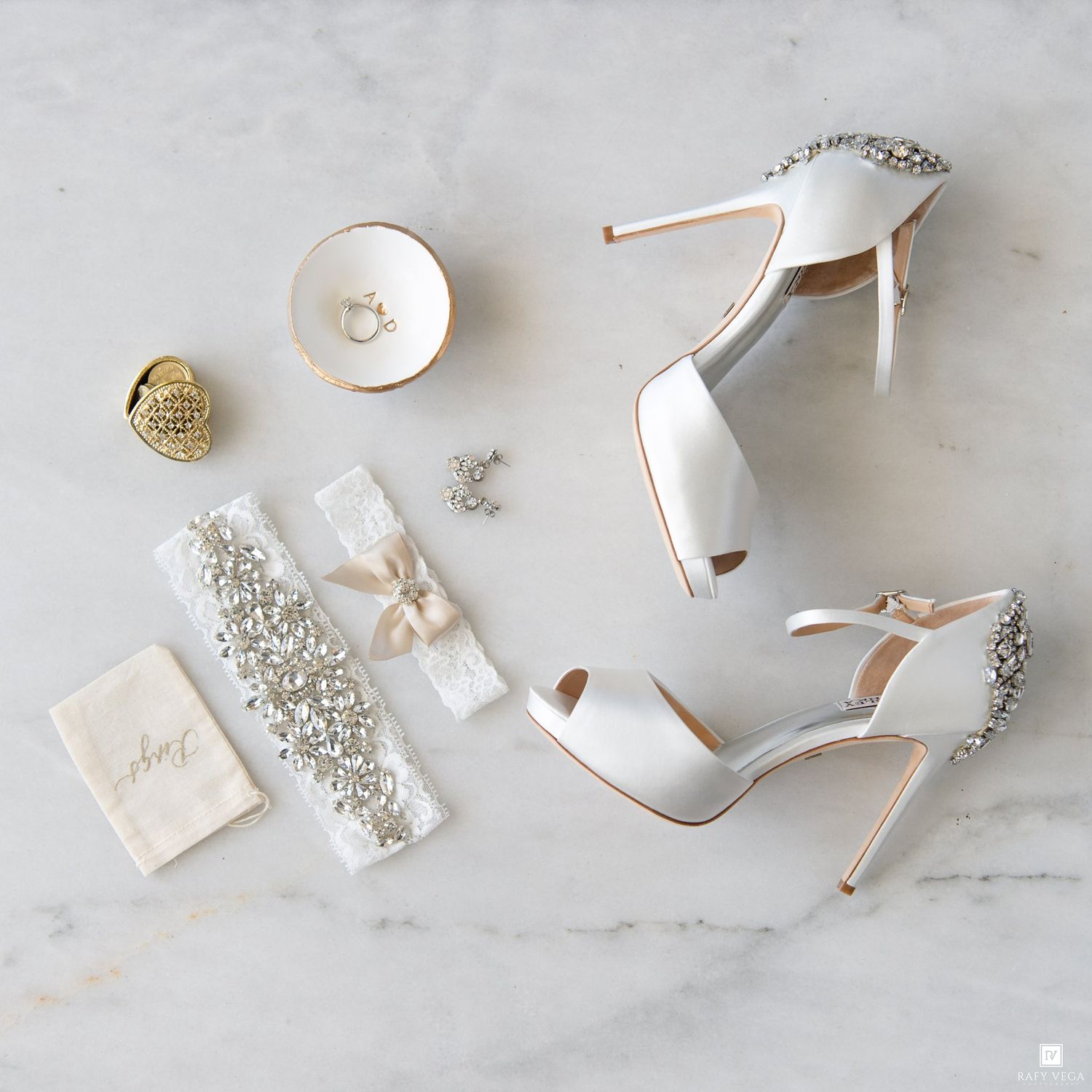 Elegant bridal accessories flat lay featuring white stiletto heels, crystal garter, rings, and jewelry on marble.