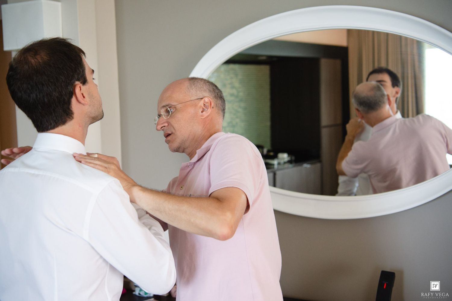 An older man helps a younger man adjust his white dress shirt collar in front of a hotel room mirror.