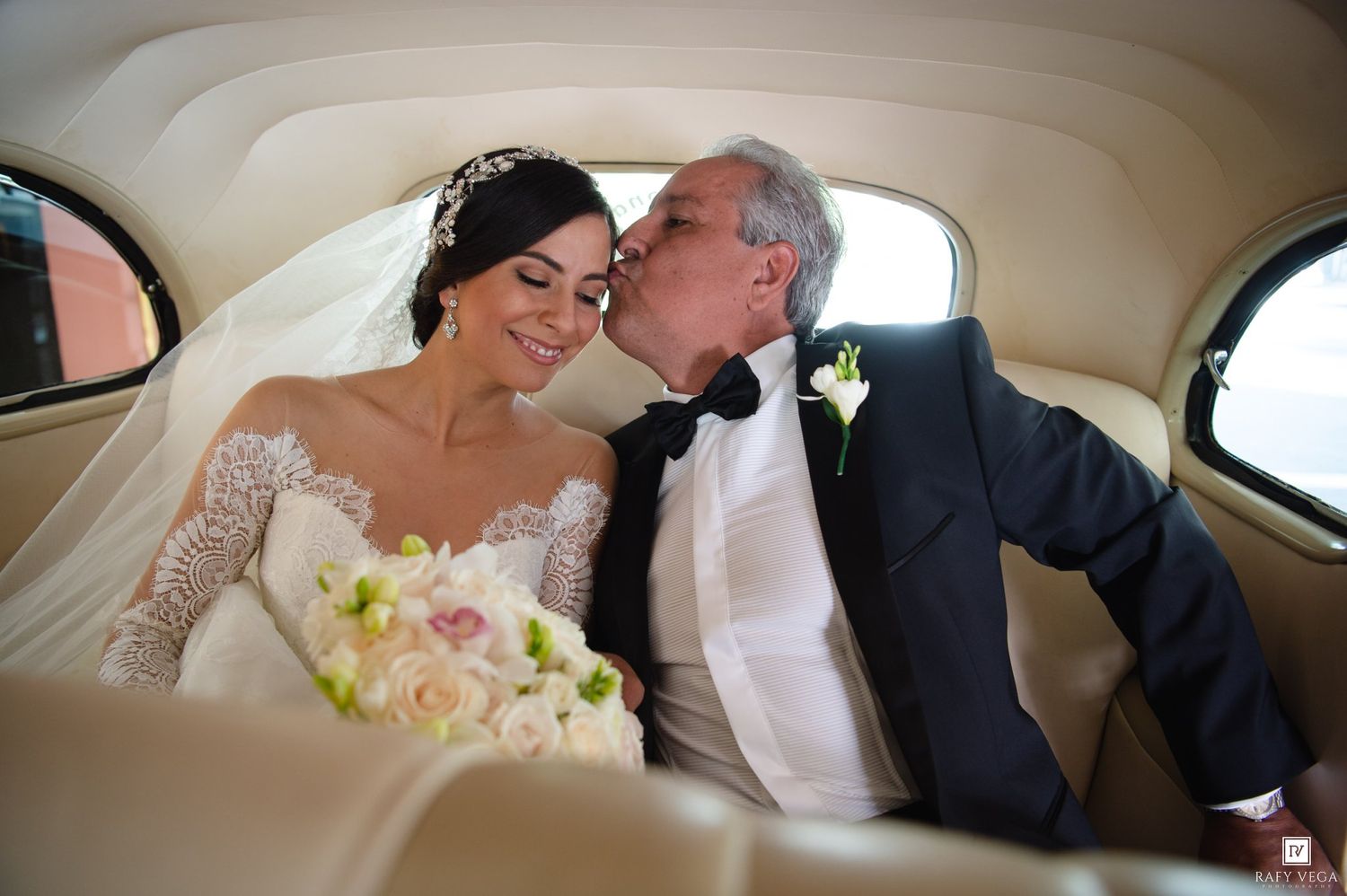 Bride in lace gown holding pink bouquet receives kiss from older man in tuxedo inside vintage car.