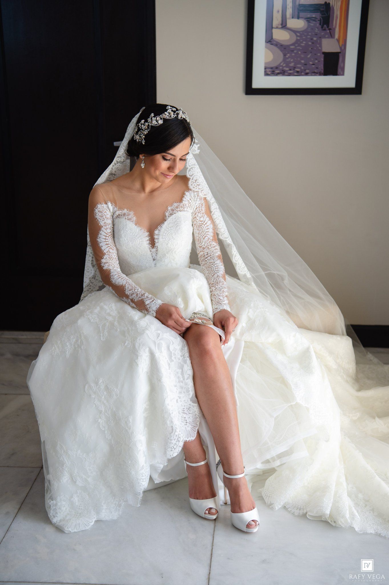 Bride in lace long-sleeve wedding gown with veil adjusting white heels while seated, elegant bridal portrait.