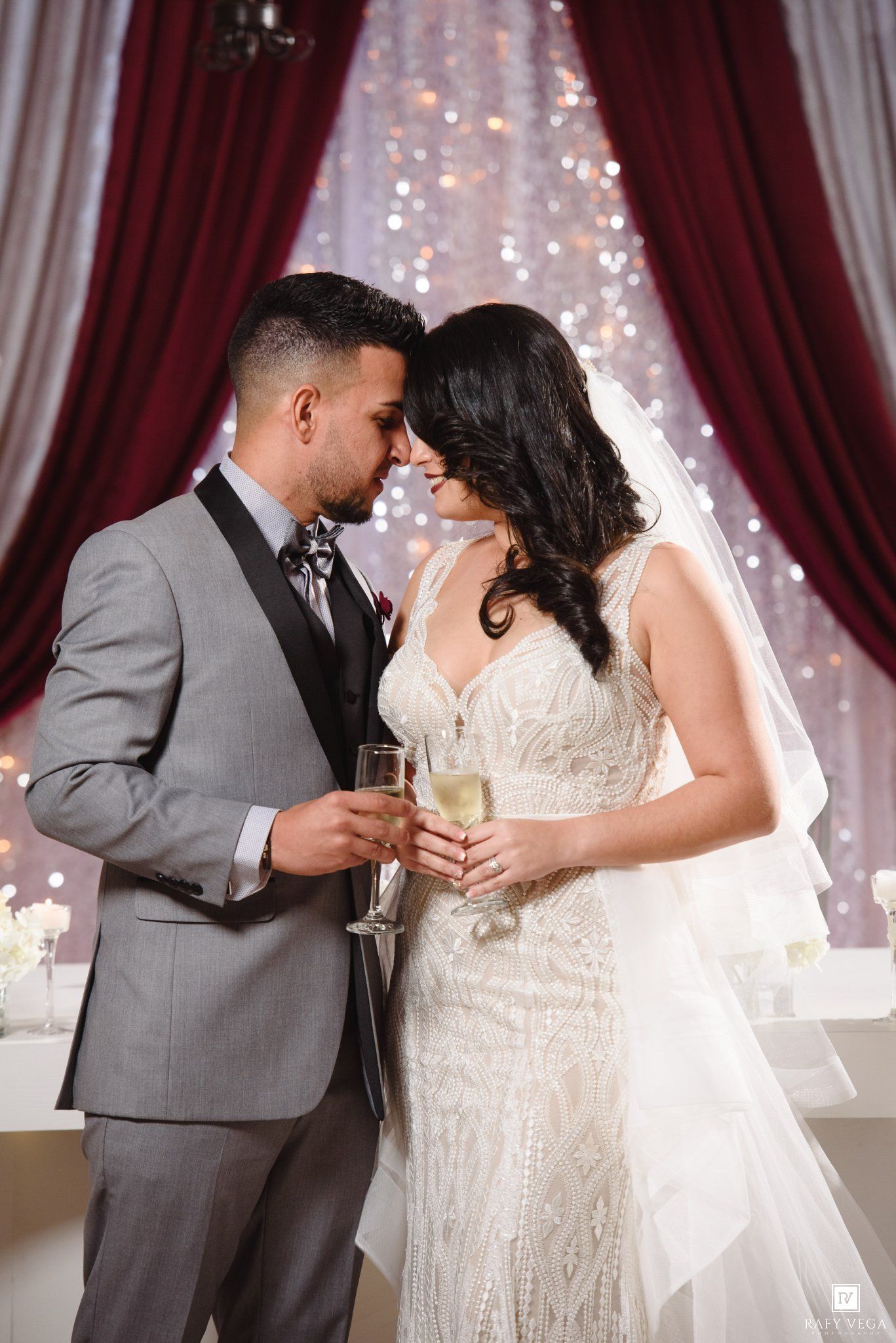 Bride and groom share an intimate moment, holding champagne glasses, dressed in wedding attire with elegant red curtain backdrop.