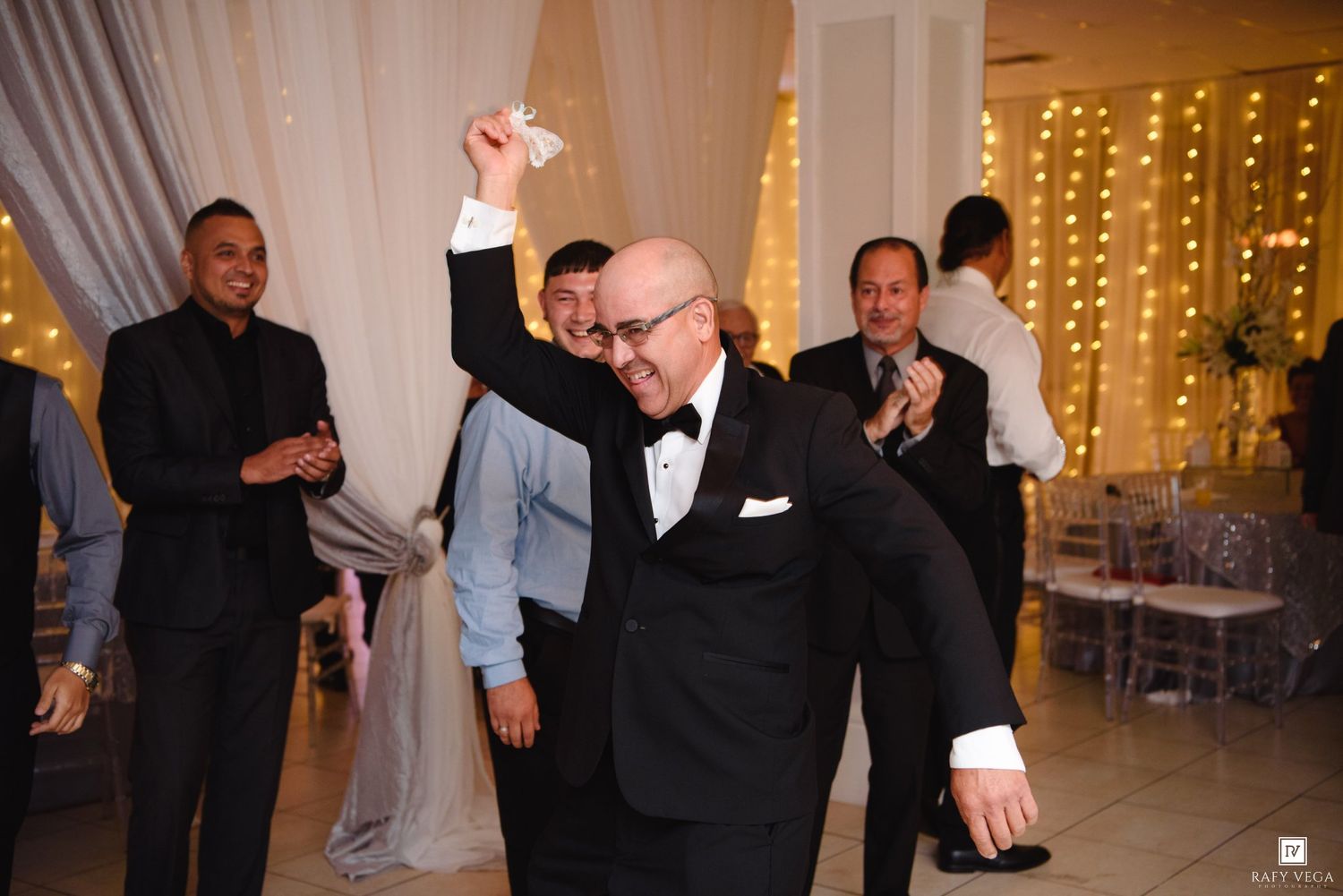 Elderly man in tuxedo dancing joyfully at wedding reception with guests applauding in background.