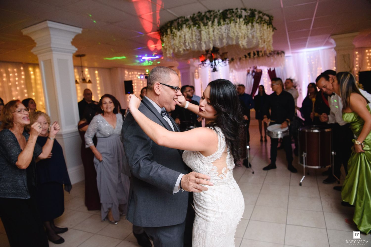Bride and groom share first dance at wedding reception surrounded by guests and floral chandelier.
