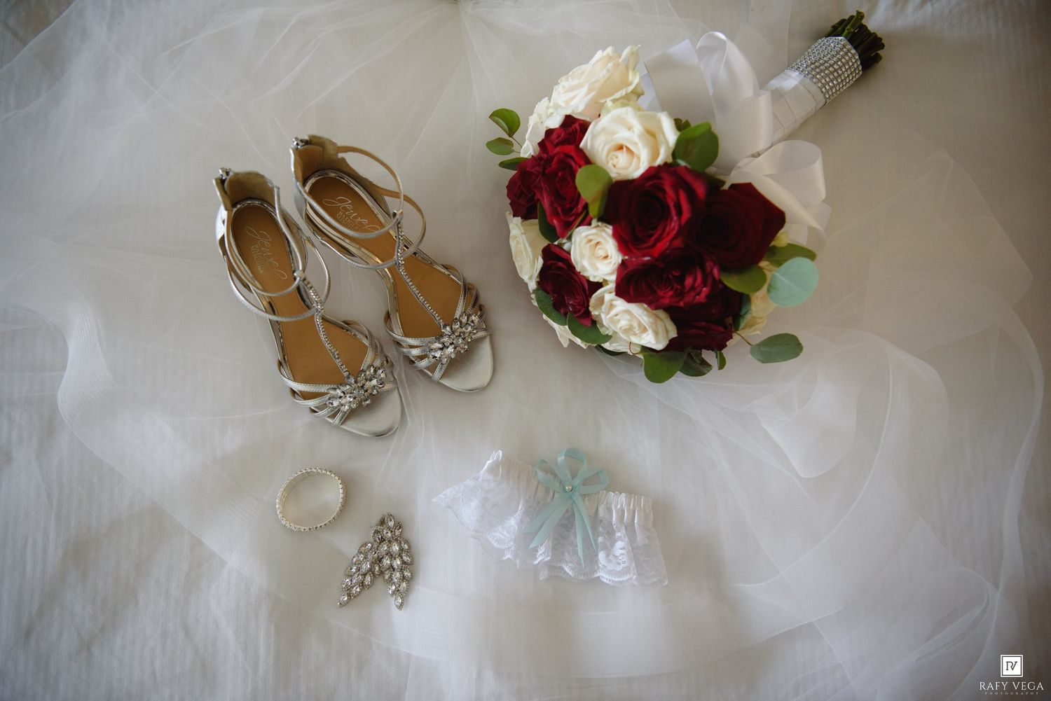 Bridal flat lay with silver strappy heels, red and white rose bouquet, garter, earrings, and ring on white tulle fabric.