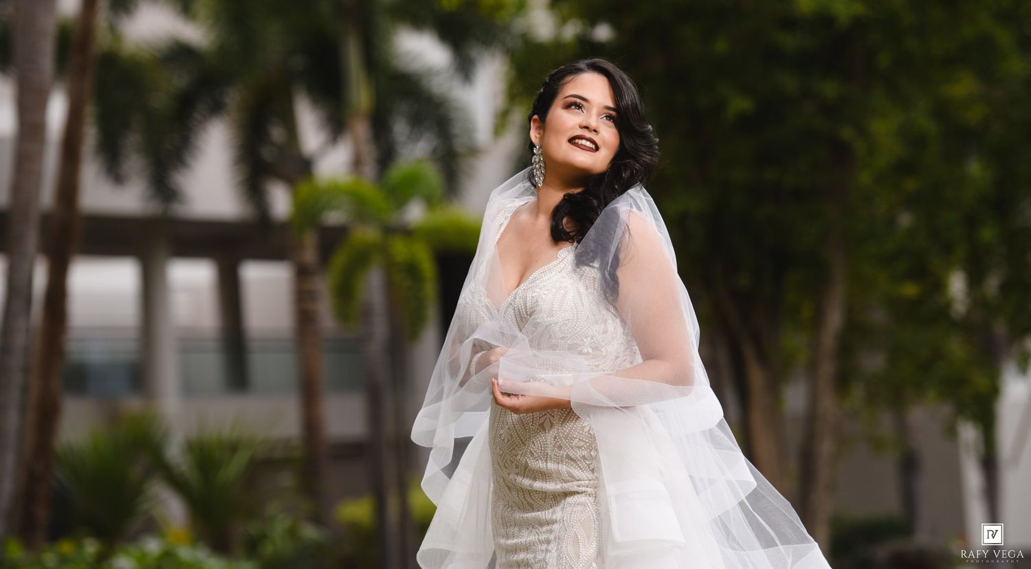 Elegant bride in a white wedding gown and veil poses outdoors with tropical palm trees in the background.
