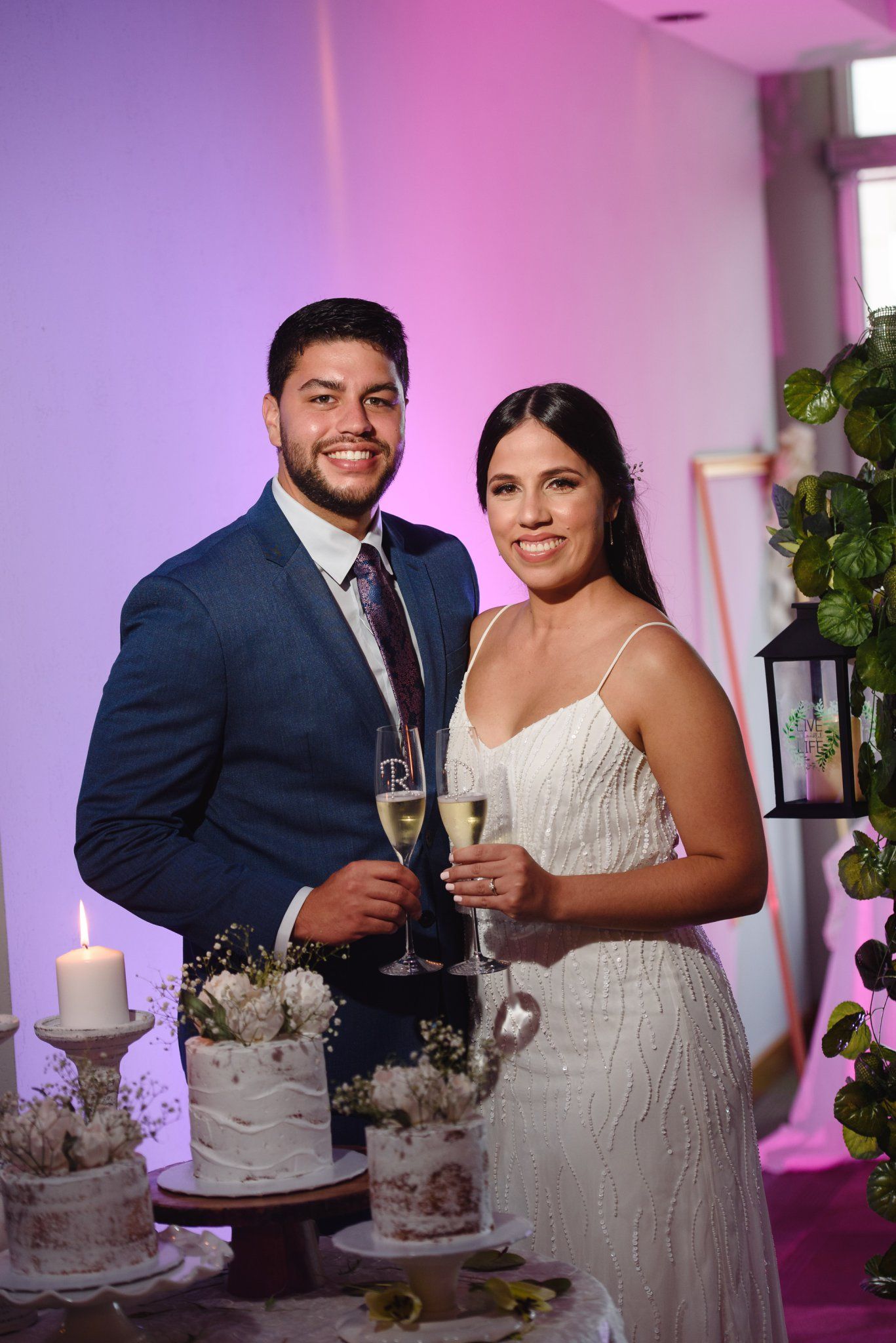 Bride and groom smiling holding champagne glasses beside wedding cake at elegant reception with purple lighting.
