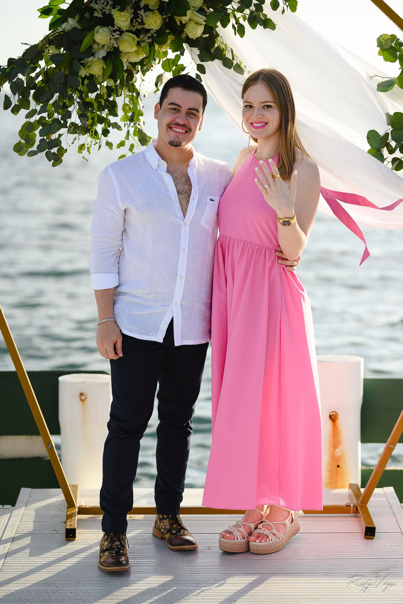 Happy couple posing on a dock, woman in pink dress showing engagement ring, man in white shirt, waterfront backdrop.