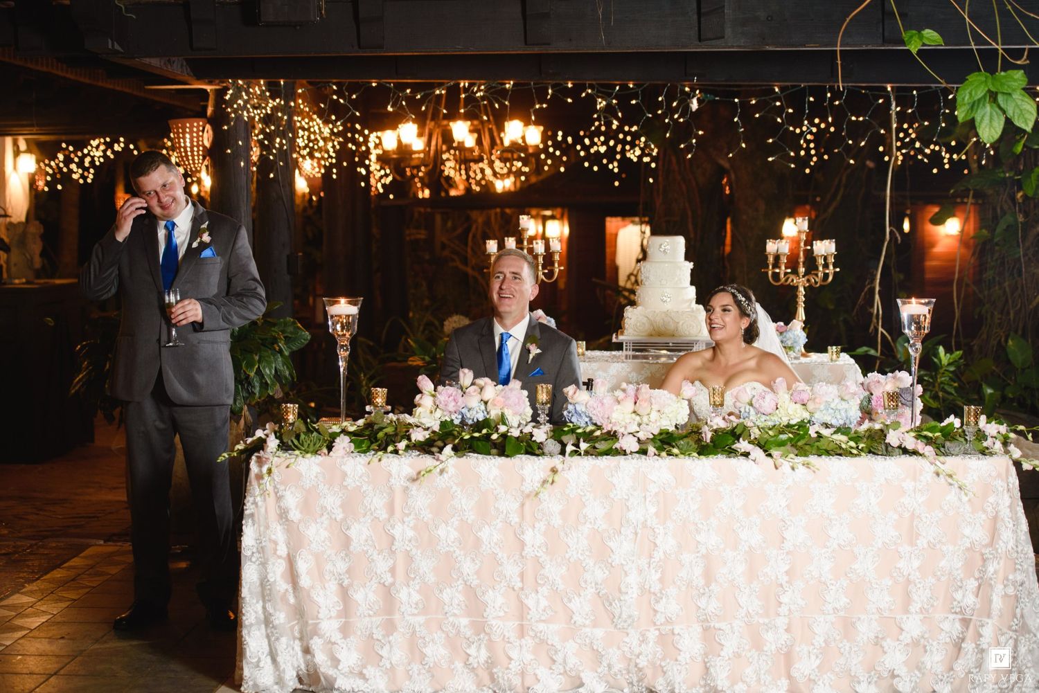 Wedding reception toast with guests at elegantly decorated head table with floral arrangements and tiered cake.