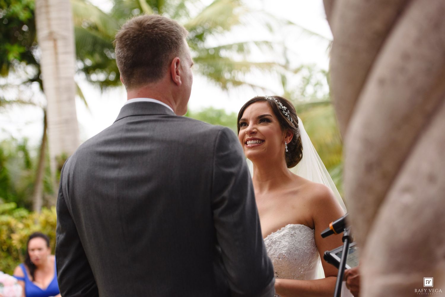 Bride smiling joyfully at groom during outdoor wedding ceremony with tropical palm trees in background.