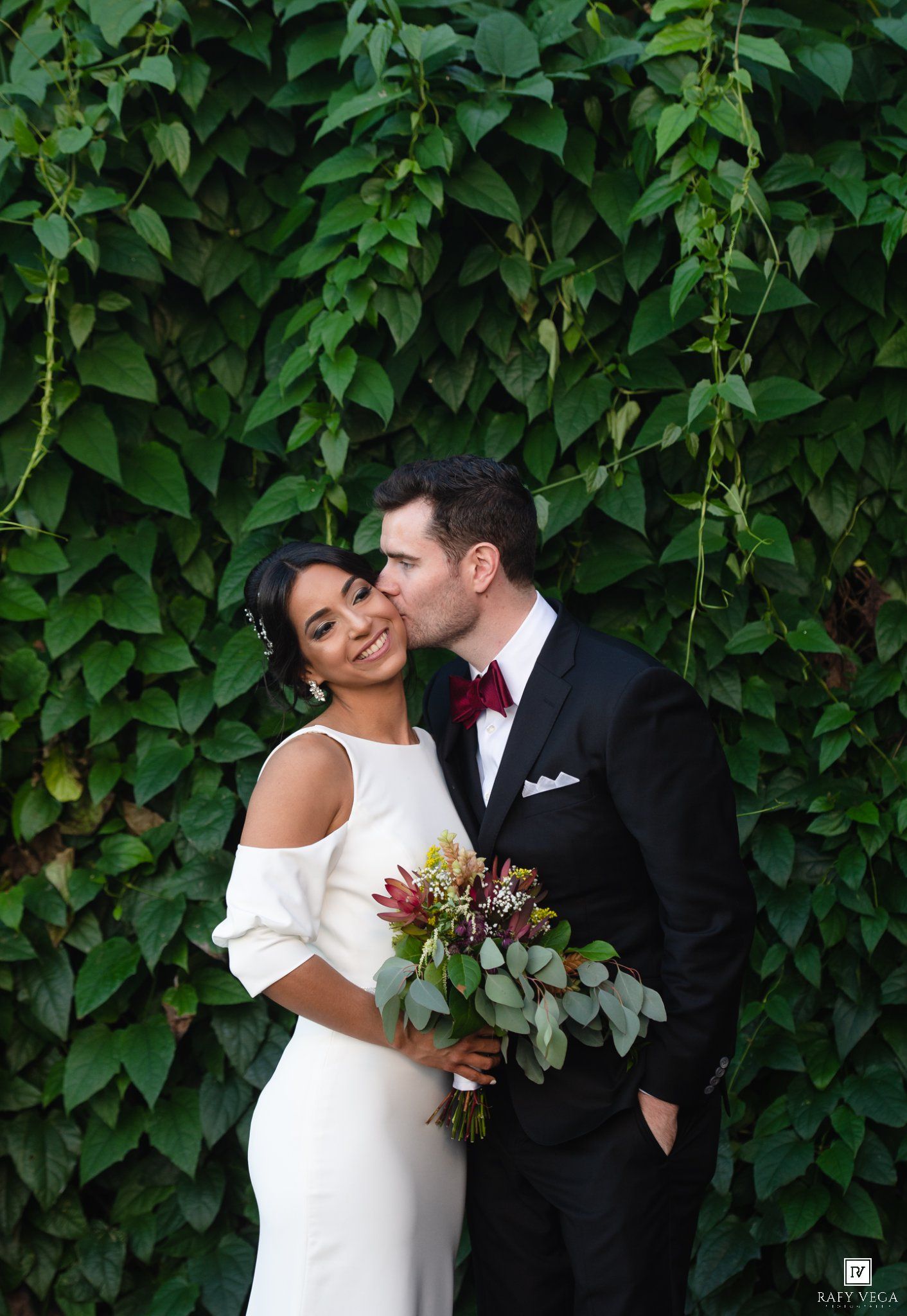 Bride and groom share a romantic kiss in front of a lush green ivy wall, bride holding a burgundy bouquet.