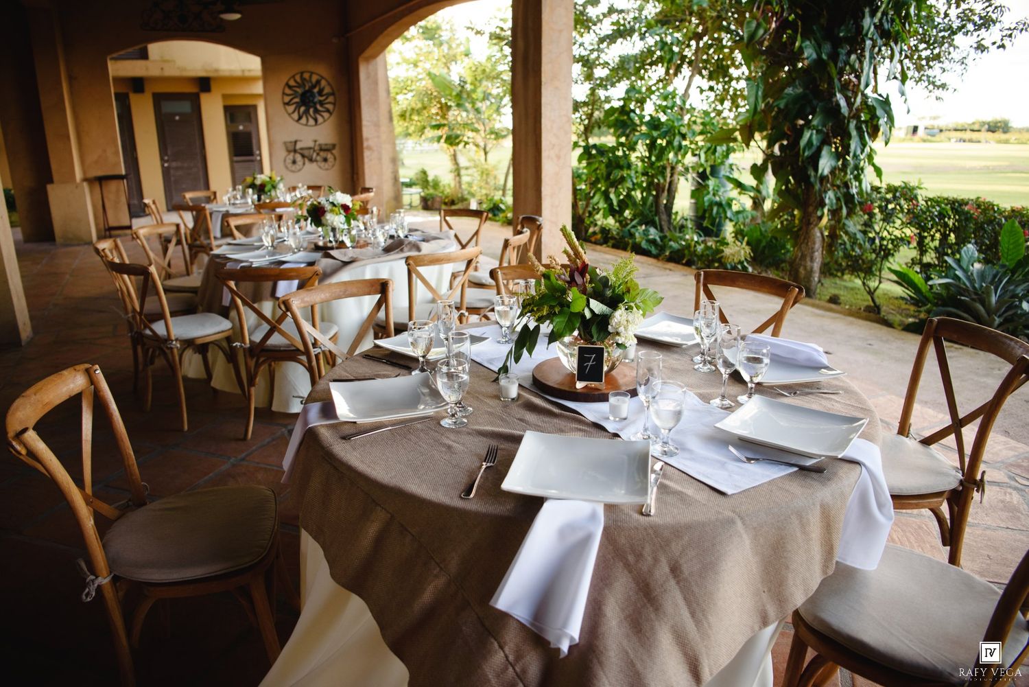 Elegant outdoor wedding reception with round tables set with burlap linens, white napkins, and wooden cross-back chairs.