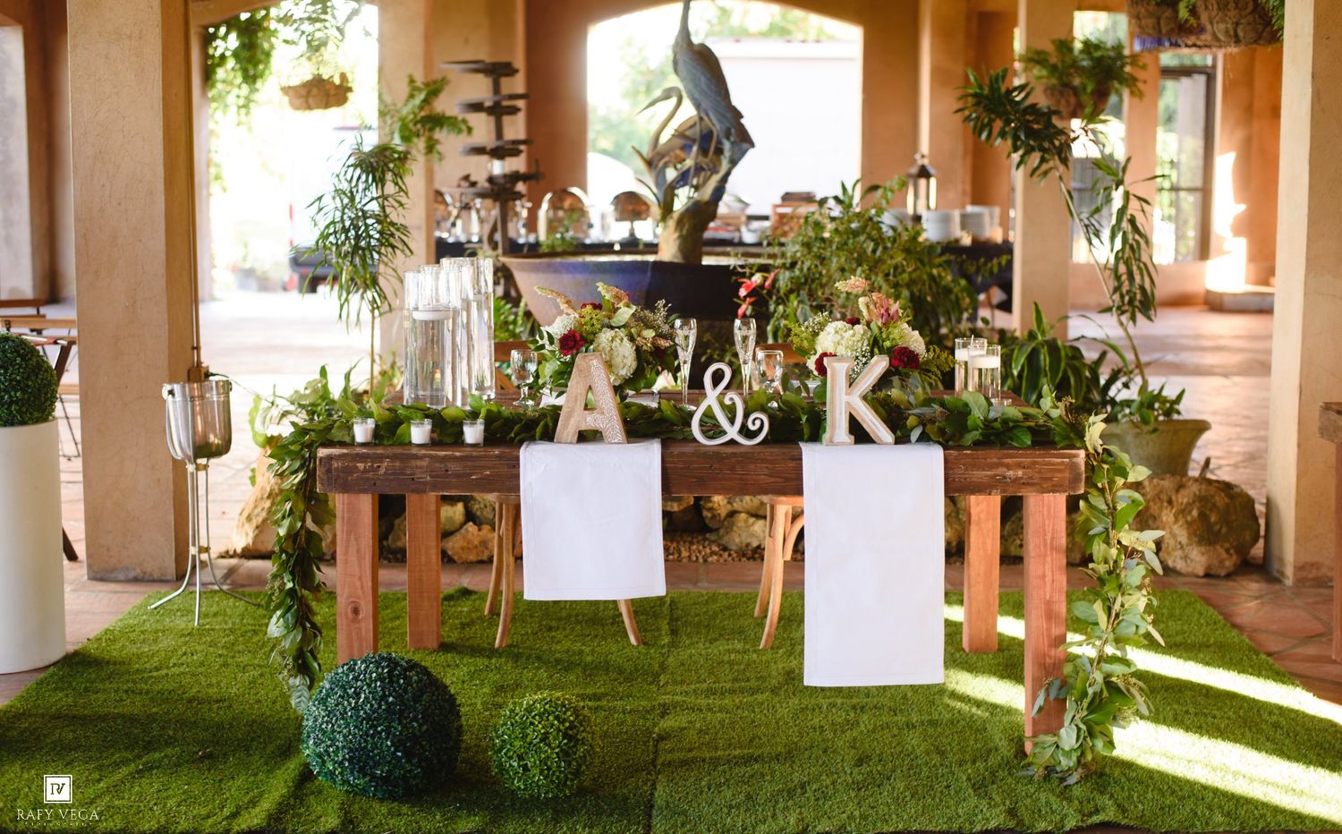 Elegant wedding sweetheart table decorated with lush greenery, wooden letters A&K, and floral arrangements on artificial grass.