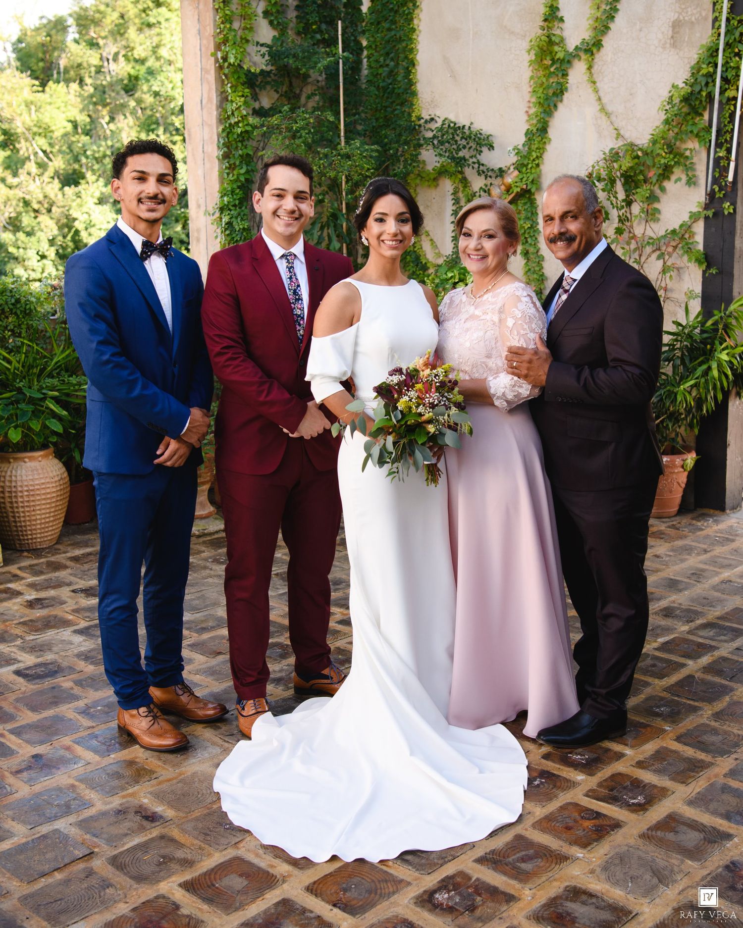 Wedding party poses outdoors with bride in white gown holding bouquet, flanked by groomsmen in blue and burgundy suits.