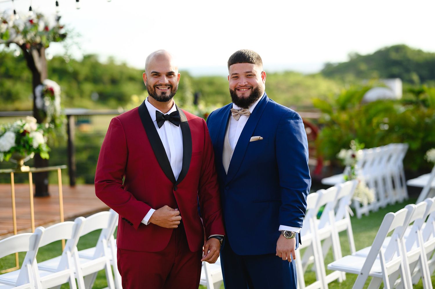 Two grooms in red and blue suits smile together at an outdoor wedding ceremony with white chairs.