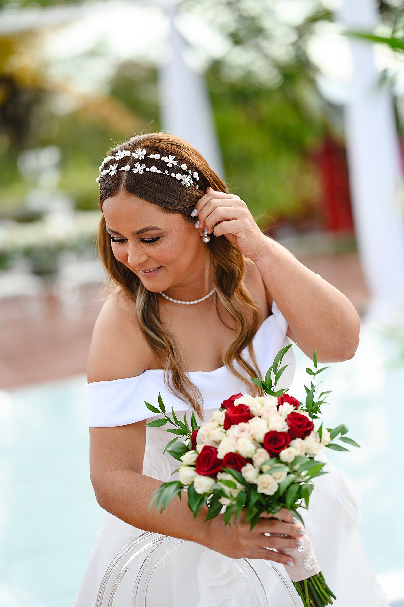 Bride in off-shoulder white gown holds red and white rose bouquet, wearing floral headpiece near a pool.