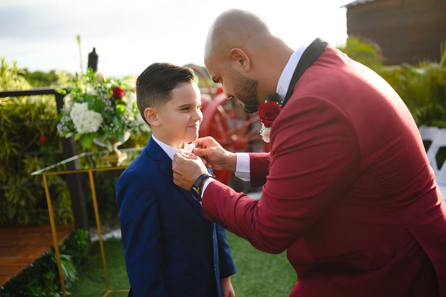 A man in a red suit adjusts a boutonniere on a young boy in a blue suit at an outdoor wedding ceremony.