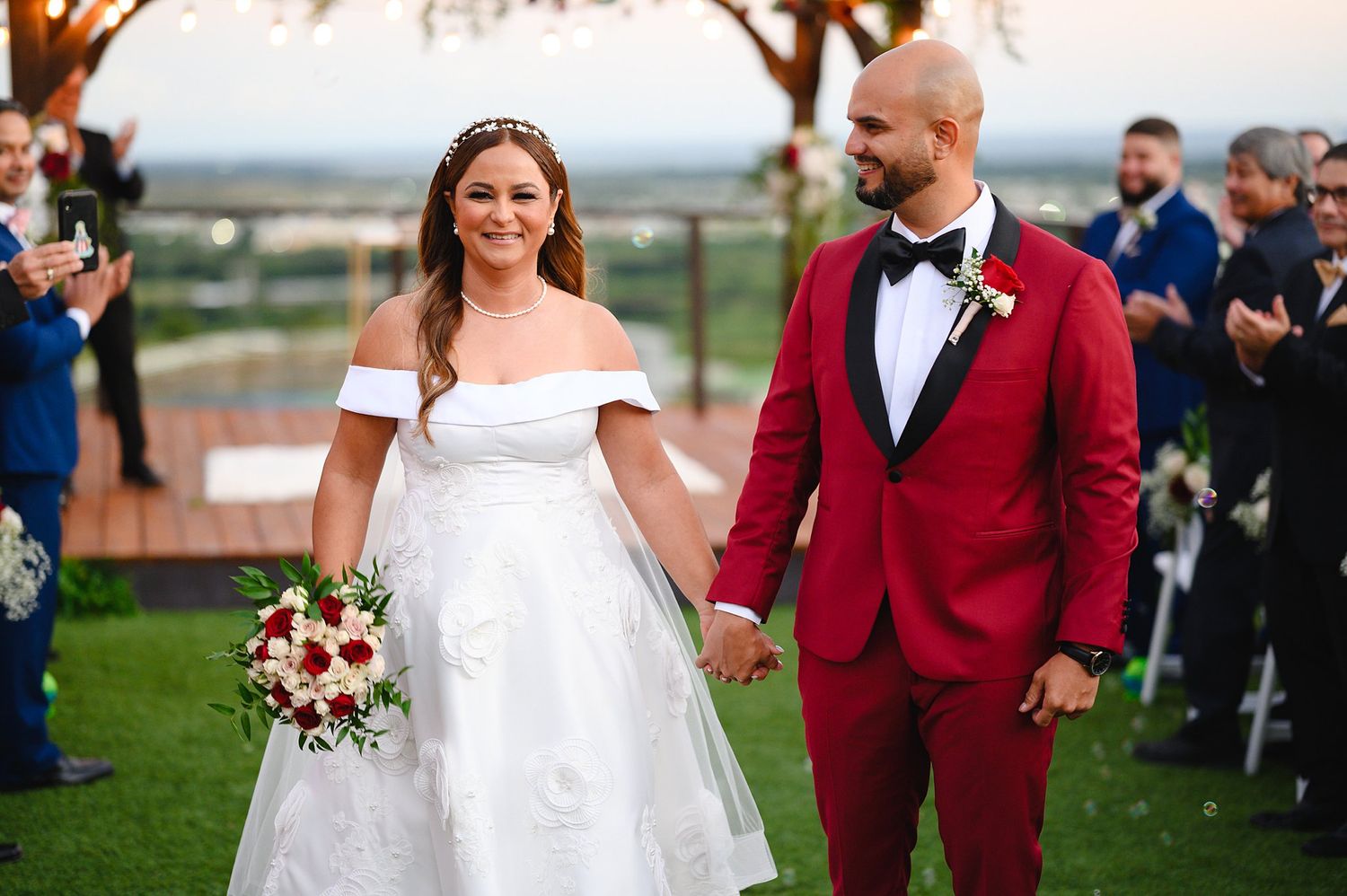Bride in white off-shoulder gown and groom in red suit hold hands during outdoor wedding ceremony.
