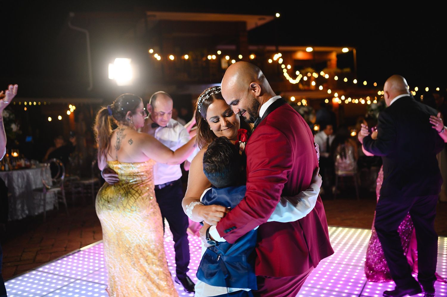 Couple embracing family members in a group hug on a lit dance floor at an outdoor wedding reception at night.