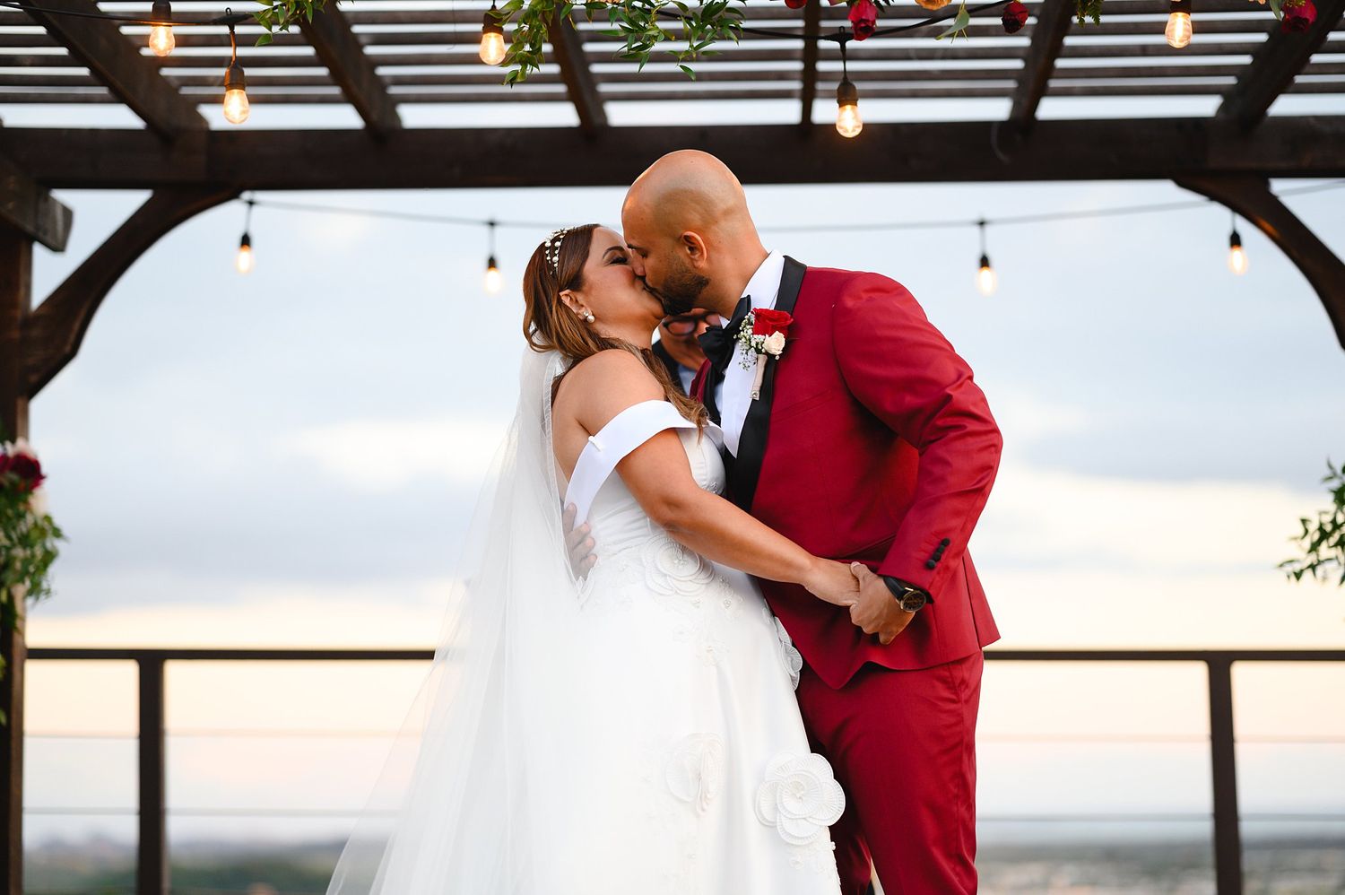 Bride in white off-shoulder gown and groom in red suit share first kiss under pergola with string lights at sunset.