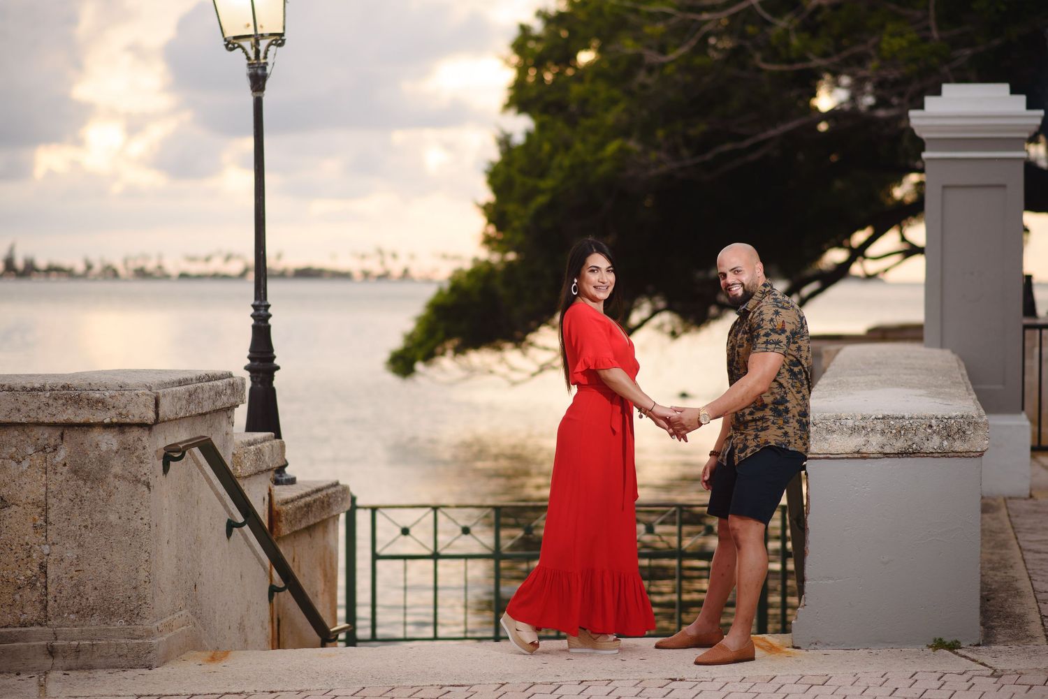 Couple holding hands on waterfront steps at sunset, woman in red dress, man in plaid shirt, lush trees and calm water behind.