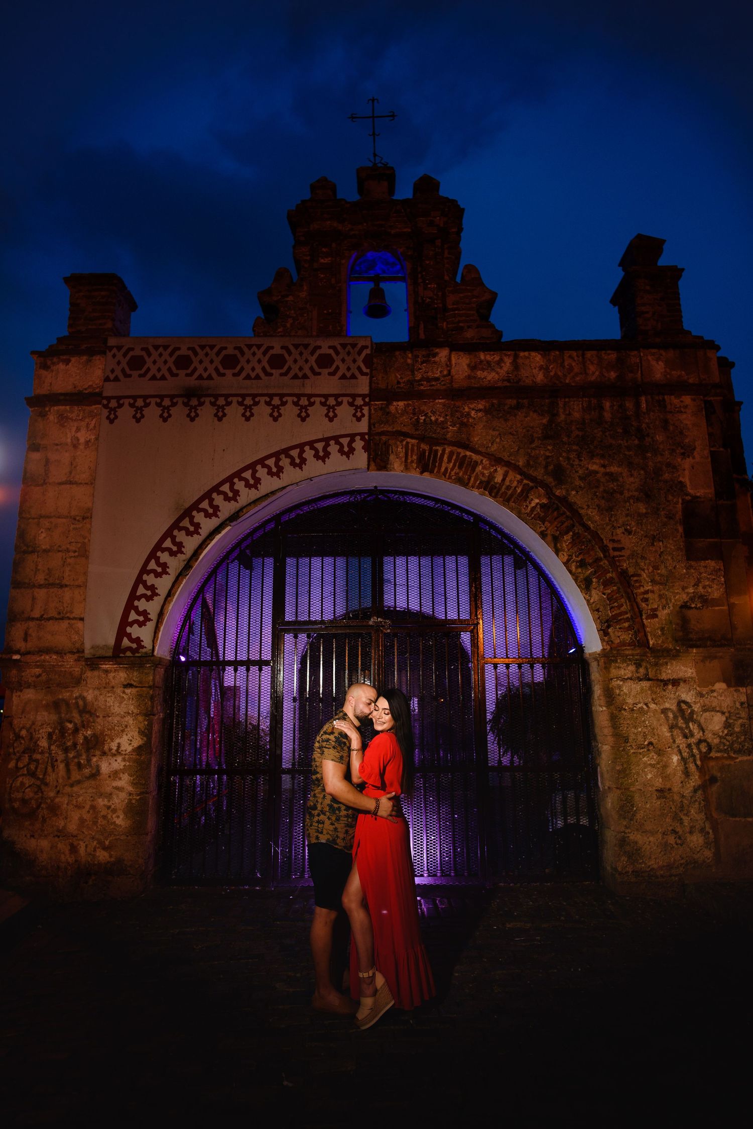 Couple embracing at night in front of an ornate stone archway gate with a bell tower and cross.