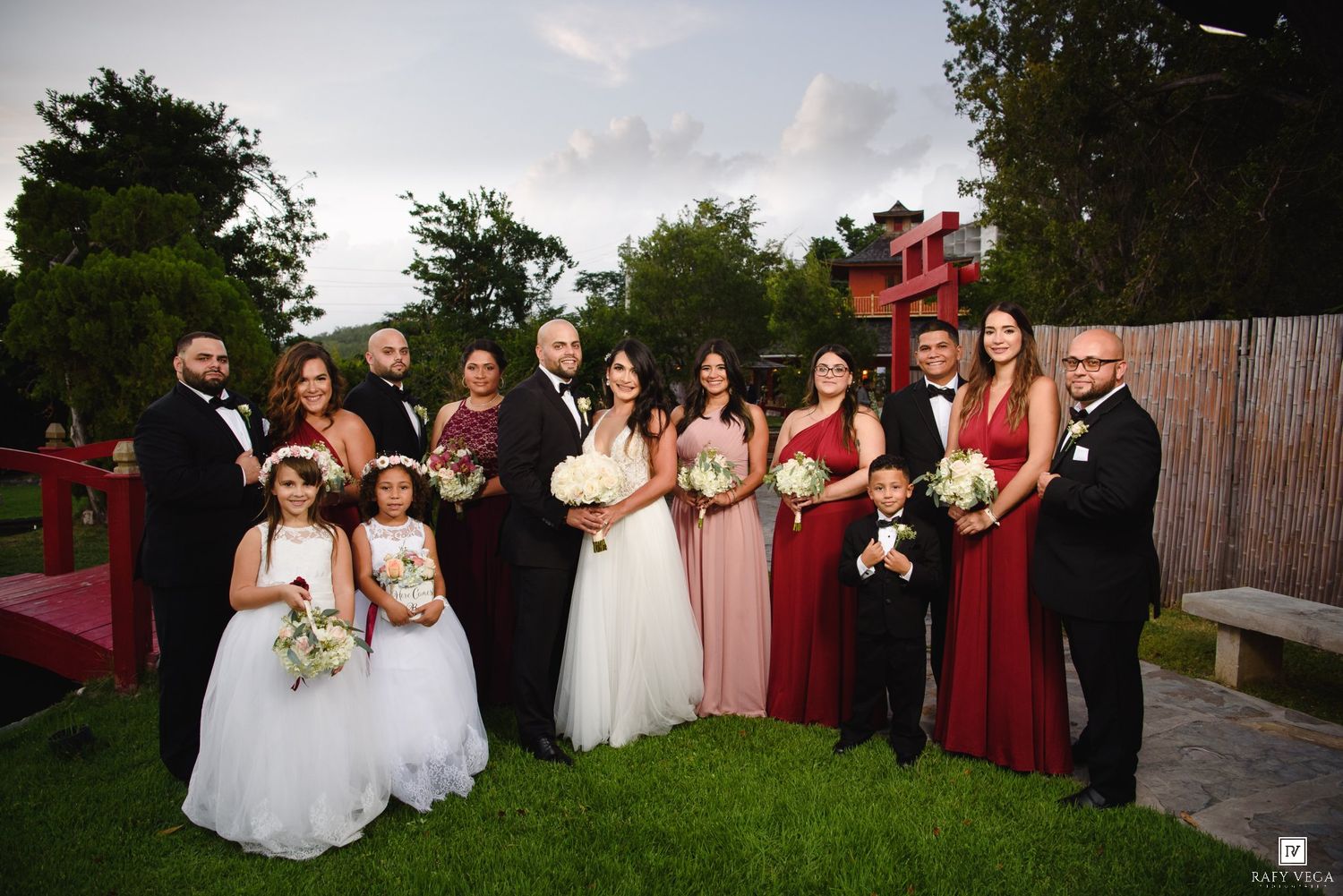 Wedding party poses outdoors in red and pink attire with bride and groom centered on lush green lawn.