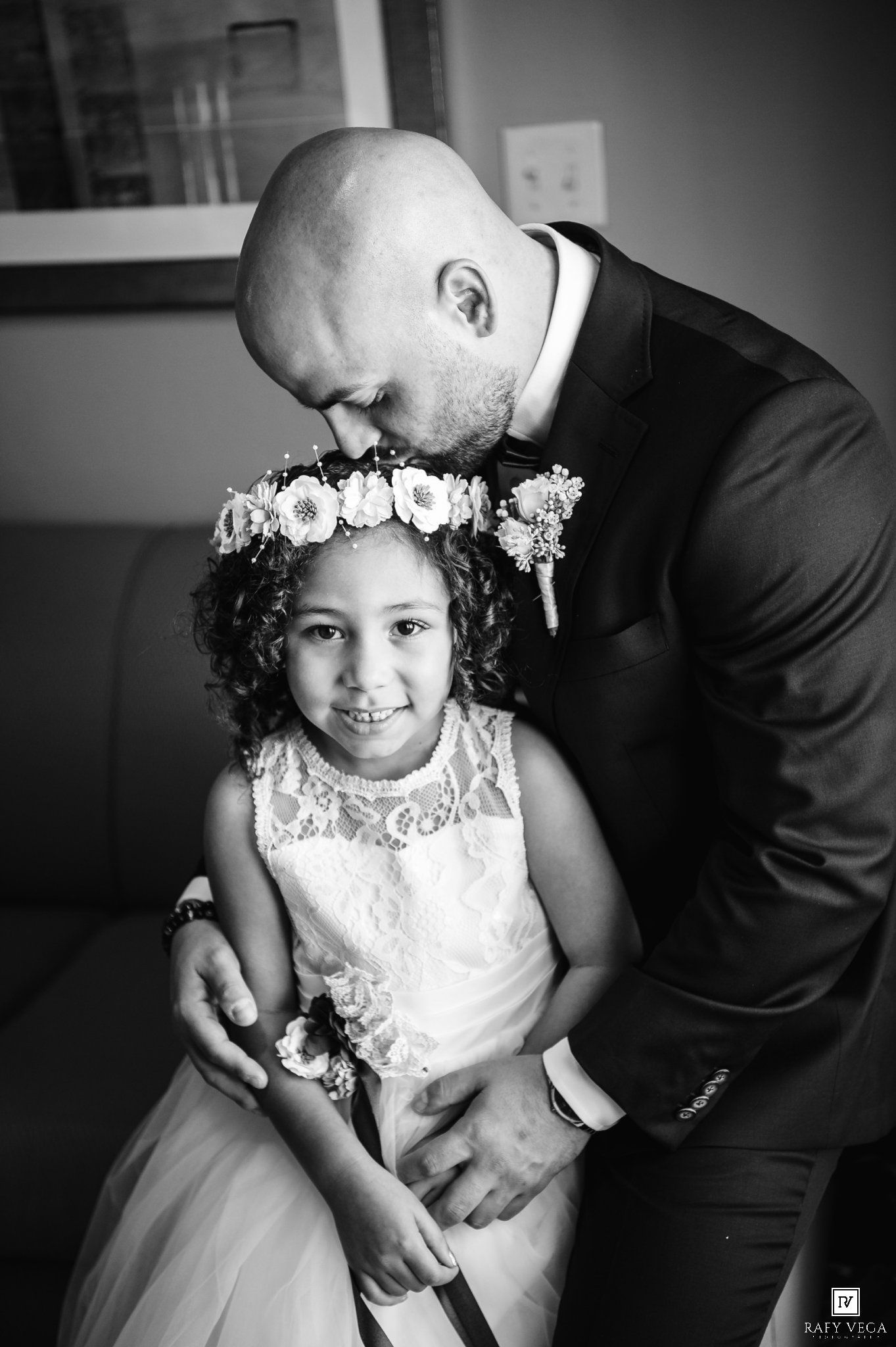 Bald groom in suit kissing flower girl wearing floral crown and white lace dress in black and white wedding photo.