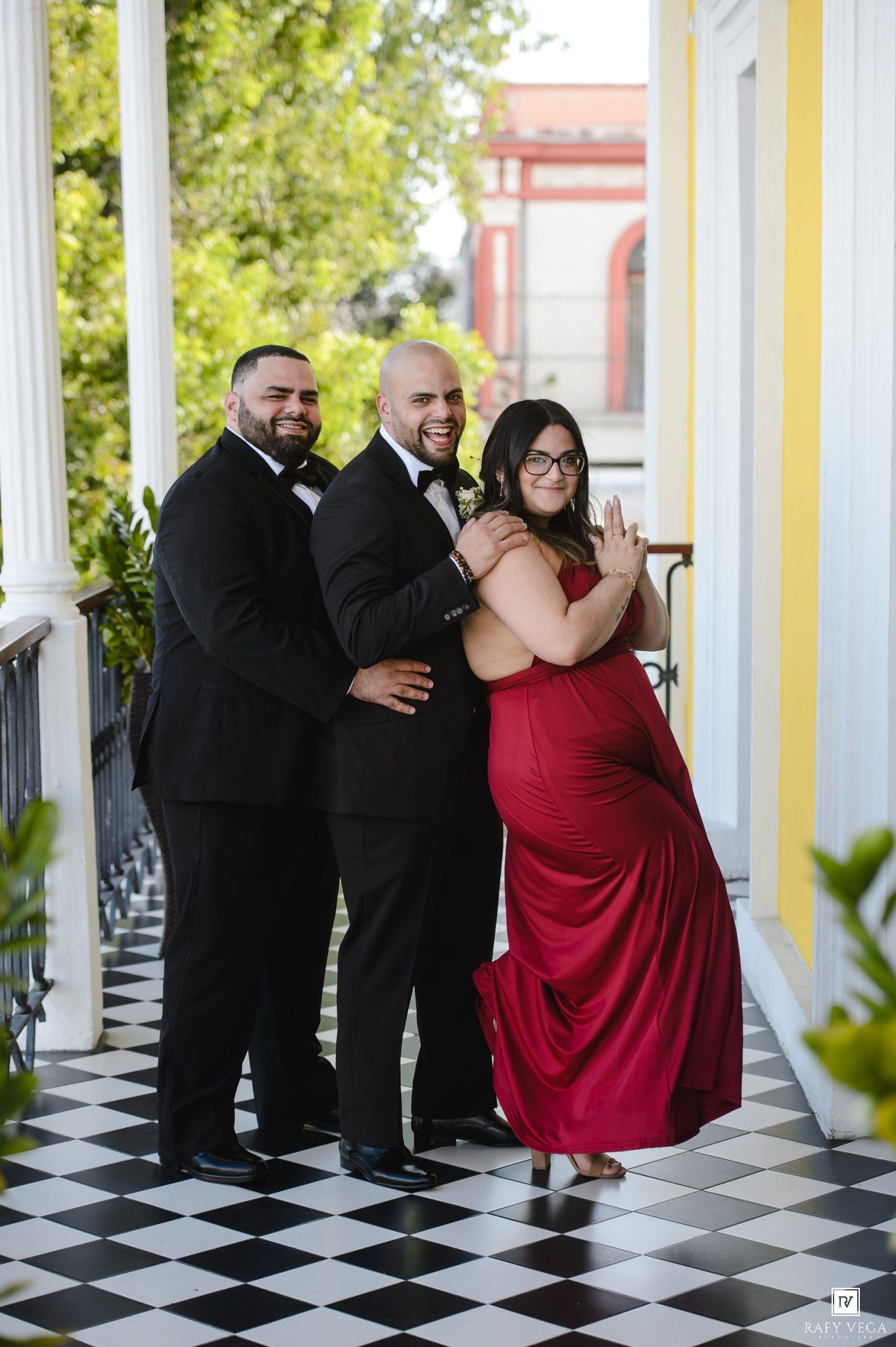 Two men in tuxedos and a woman in a red gown pose playfully on a checkered balcony floor.