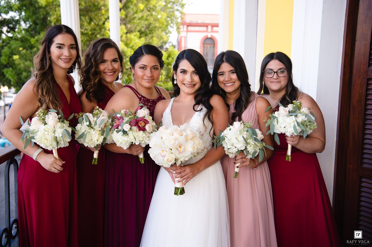 Bride in white gown poses with five bridesmaids in burgundy and blush dresses holding white floral bouquets outdoors.