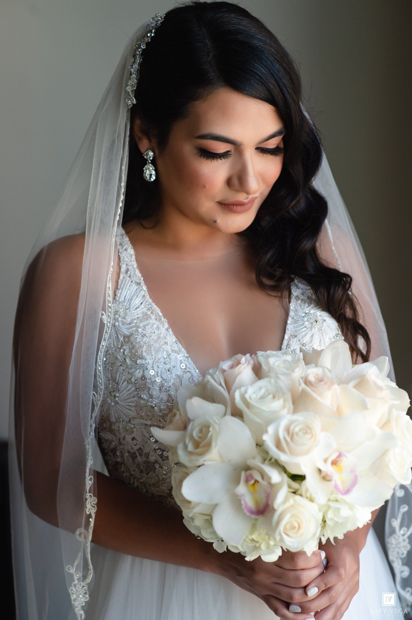 Bride in lace wedding gown and veil holds white rose and orchid bouquet, looking down elegantly.