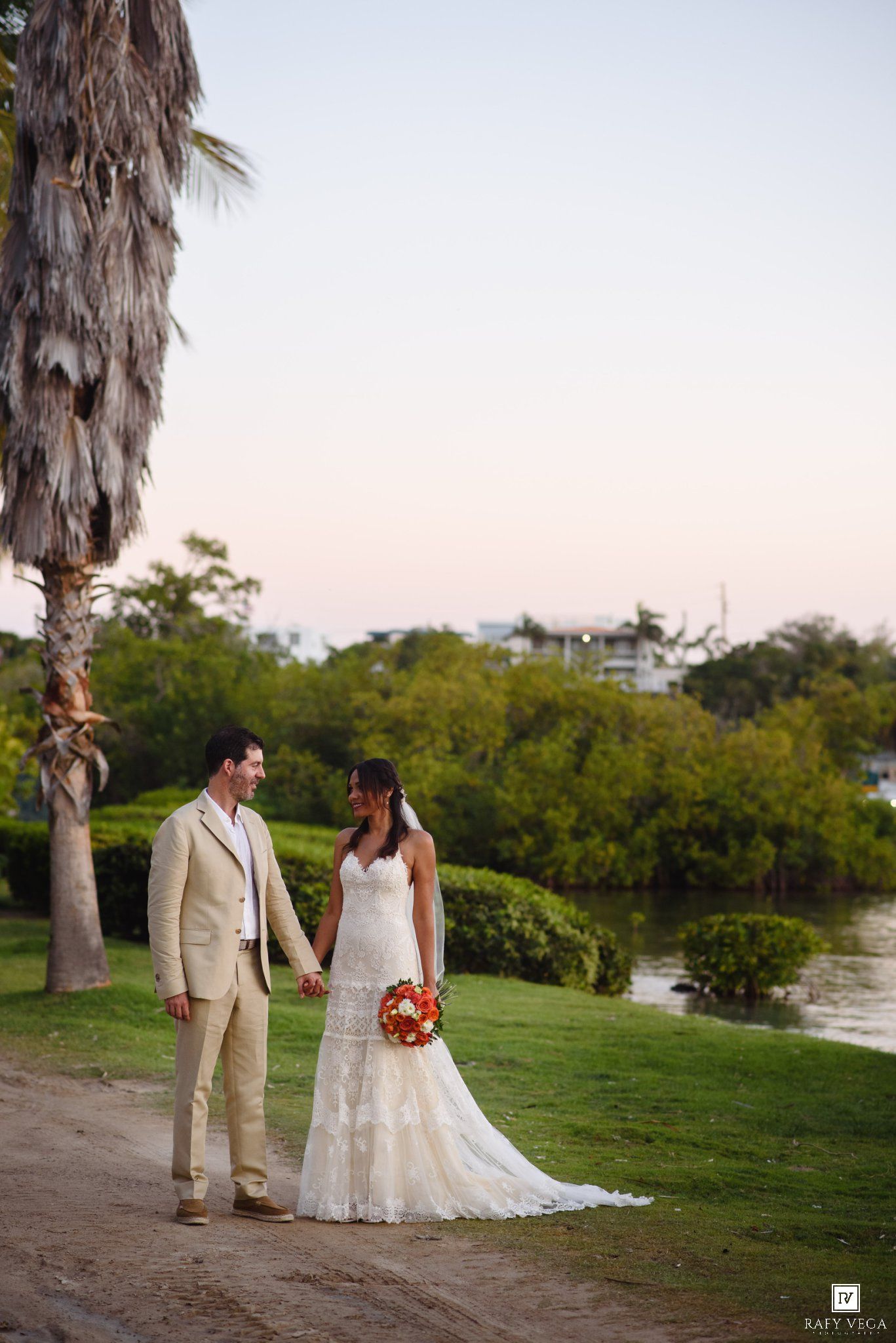 Bride and groom hold hands at sunset near a tropical waterway, she in lace gown with red bouquet, he in tan suit.
