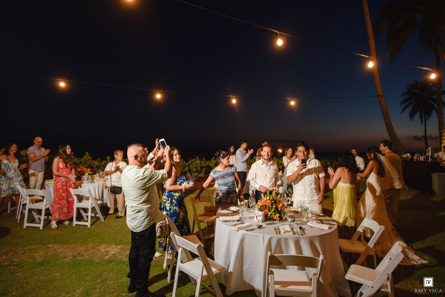 Outdoor evening wedding reception with guests dancing under string lights near the ocean at night.