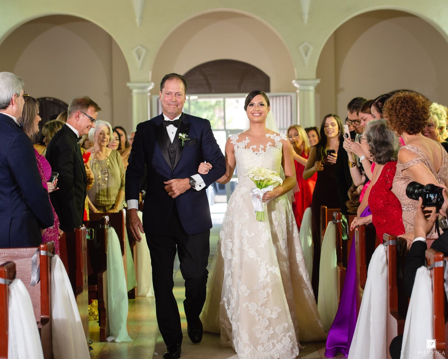 Bride in lace gown walks down church aisle with escort as wedding guests watch and photograph the ceremony.