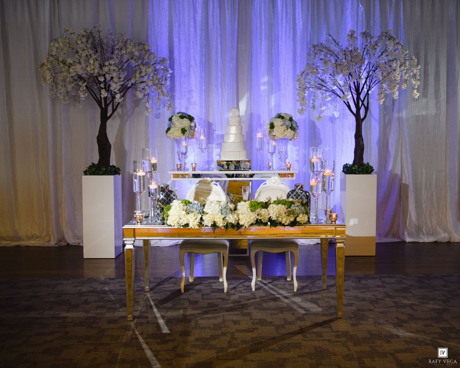 Elegant wedding sweetheart table with white floral arrangements, tiered cake, candles, and cherry blossom trees against draped backdrop.