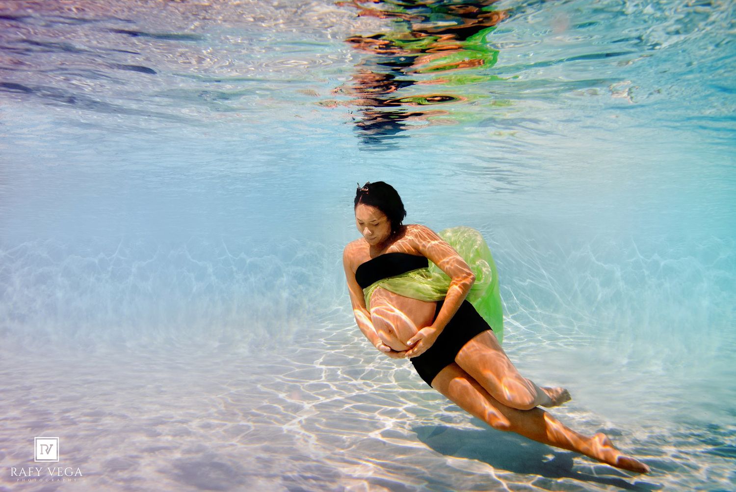 Pregnant woman in black swimsuit poses gracefully underwater in a clear blue pool, cradling her baby bump.