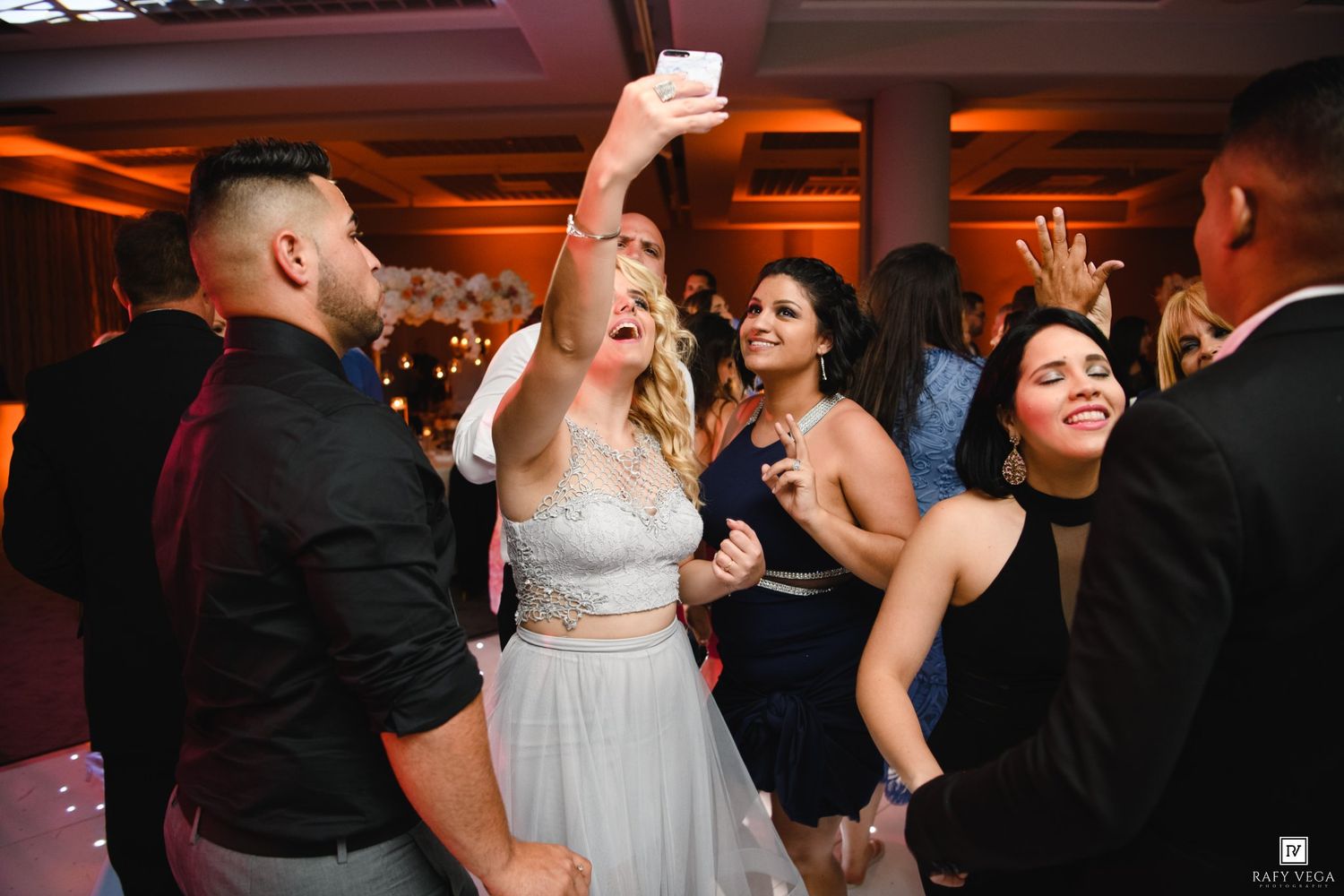 Bride takes a selfie with guests while dancing at a wedding reception in an elegant ballroom venue.
