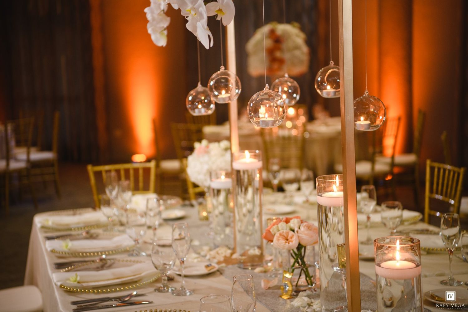 Elegant wedding reception table with gold candelabras, hanging glass orbs, candles, and blush floral centerpieces in warm amber lighting.
