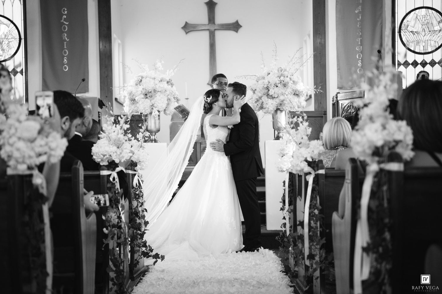 Black and white wedding photo of bride and groom kissing at church altar adorned with white floral arrangements.