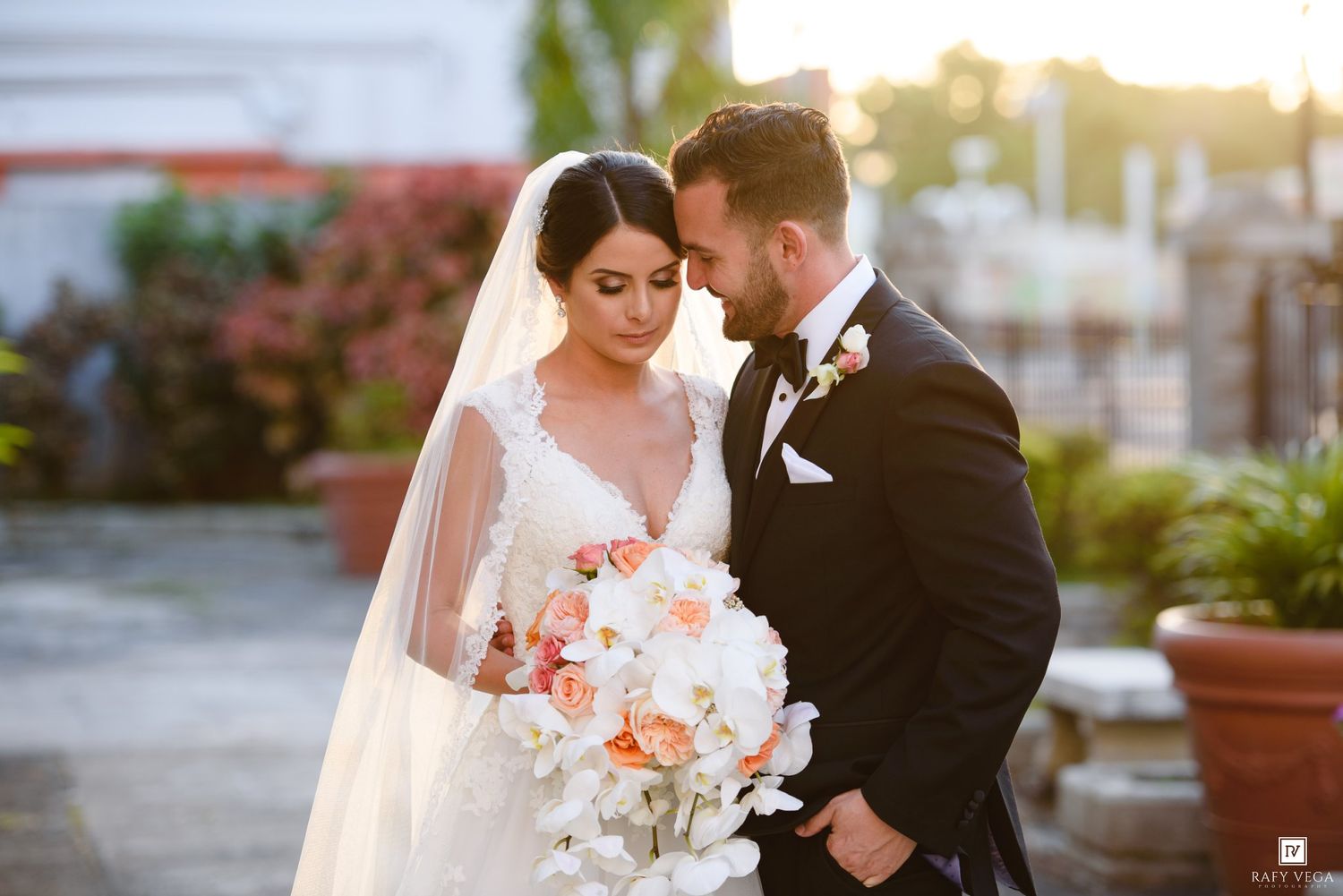 Bride and groom share an intimate moment, she holds white and peach bouquet, golden sunset backdrop.
