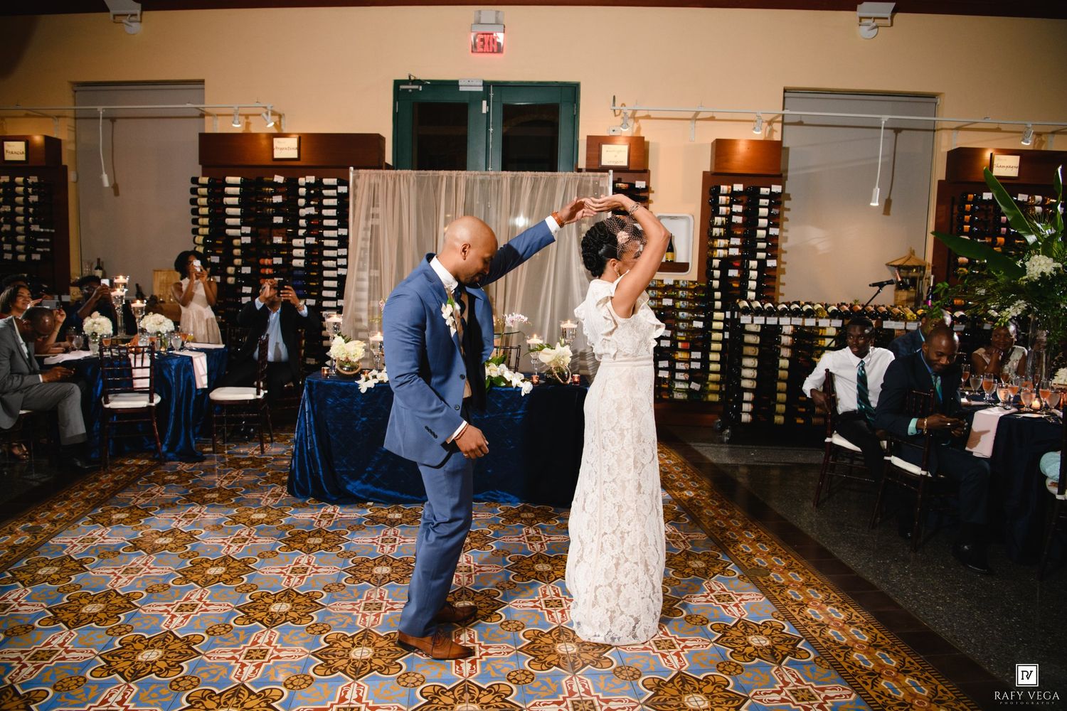 Couple shares first dance at wedding reception, bride in white gown, groom in blue suit, guests seated at decorated tables.